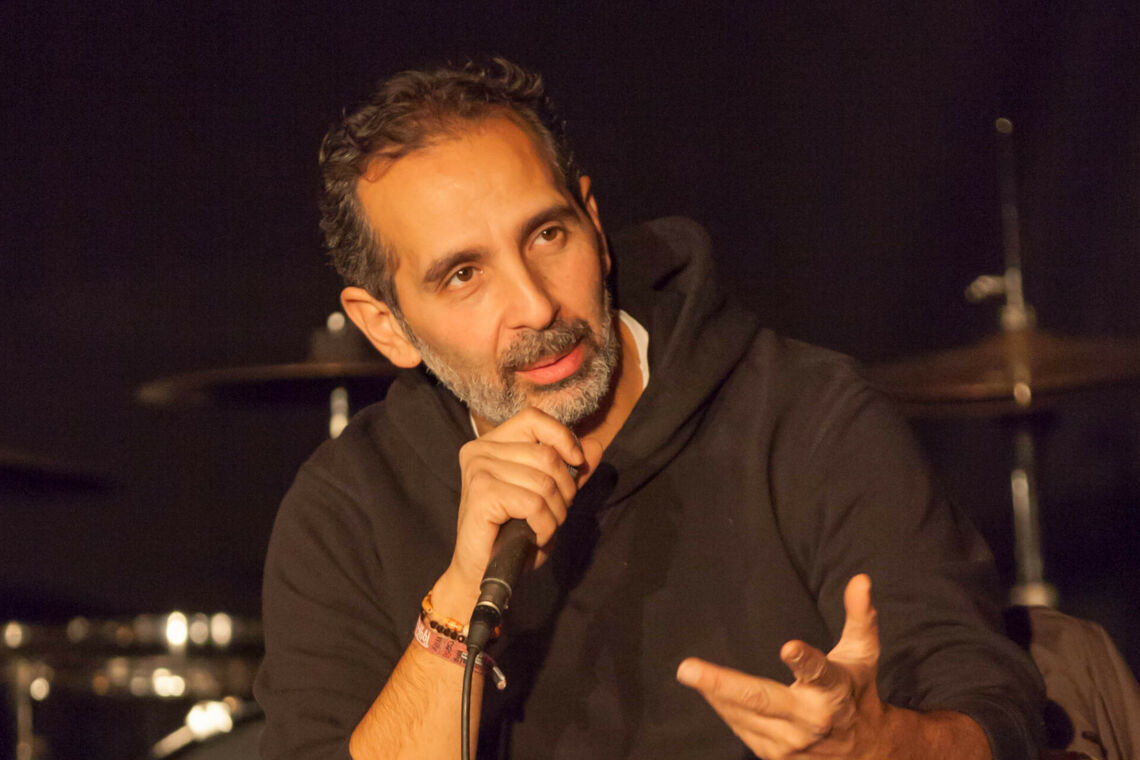 Kutlu Yurtseven spricht im schwarzen Hoodie auf einer Bühne sitzend in ein Mikrofon