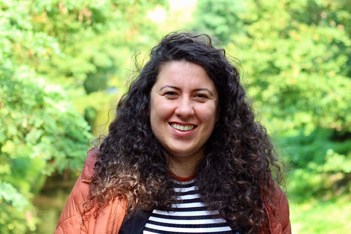 Lehrerin und Autorin Nadire Biskin schaut lächelnd in die Kamera. Sie steht draußen vor grünen Bäumen und trägt ein schwarz-weiß gestreiftes Oberteil und eine orangefarbene Jacke.