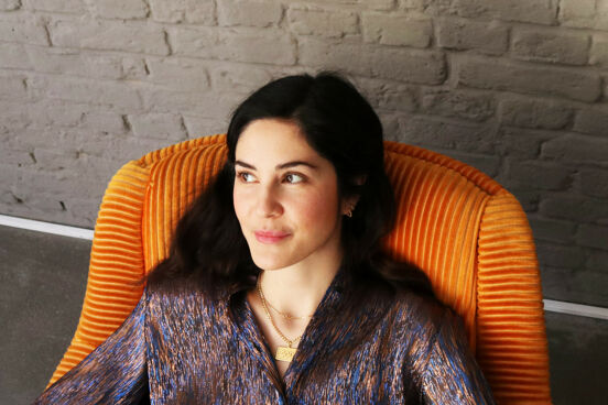 Yelda Türkmen trägt eine dunkle Bluse und sitzt in einem orangefarbenenen Sessel. Sie guckt zur Seite und lächelt.