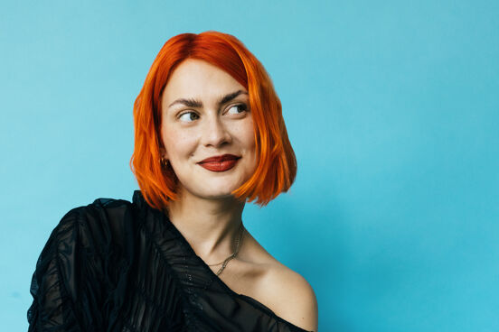 Alli Neumann trägt rote Haare, lächelt verschmitzt mit Blick nach oben linke vor hellblauem Hintergrund