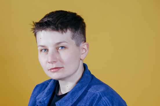 Porträtbild von Queermed-Gründer*in Sara Grzybek trägt ein blaues Jeanshemd, den Kopf leicht zur Seite geneigt vor gelbem Hintergrund.