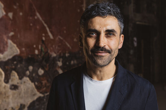 Portraitbild von Yousef Hammoudah, Executive Director von Fotografiska Berlin. Yousef trägt ein weißes Shirt und schwarzes Jacket und steht vor einer alten bröckligen Wand.