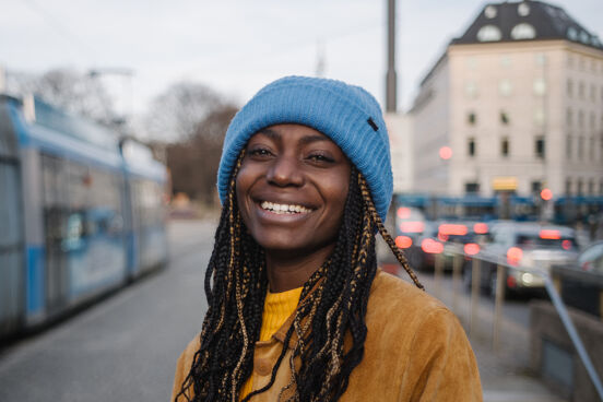 Christiana Bukalo trägt eine hellblaue Wollmütze und eine beigefarbene Jacke über einem gelben Pulli und lächelt in die Kamera.