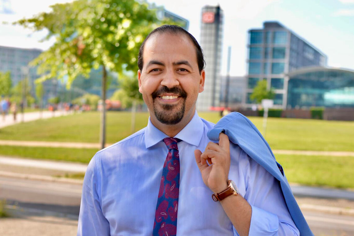 Mohammed El Ouahhabi trägt ein hellblaues Hemd, eine rotblaue Krawartte und ein blaues Sakko über der chulter. Die Sonne scheint und im Hintergrund ist der Berliner Hauptbahnhof zu sehen