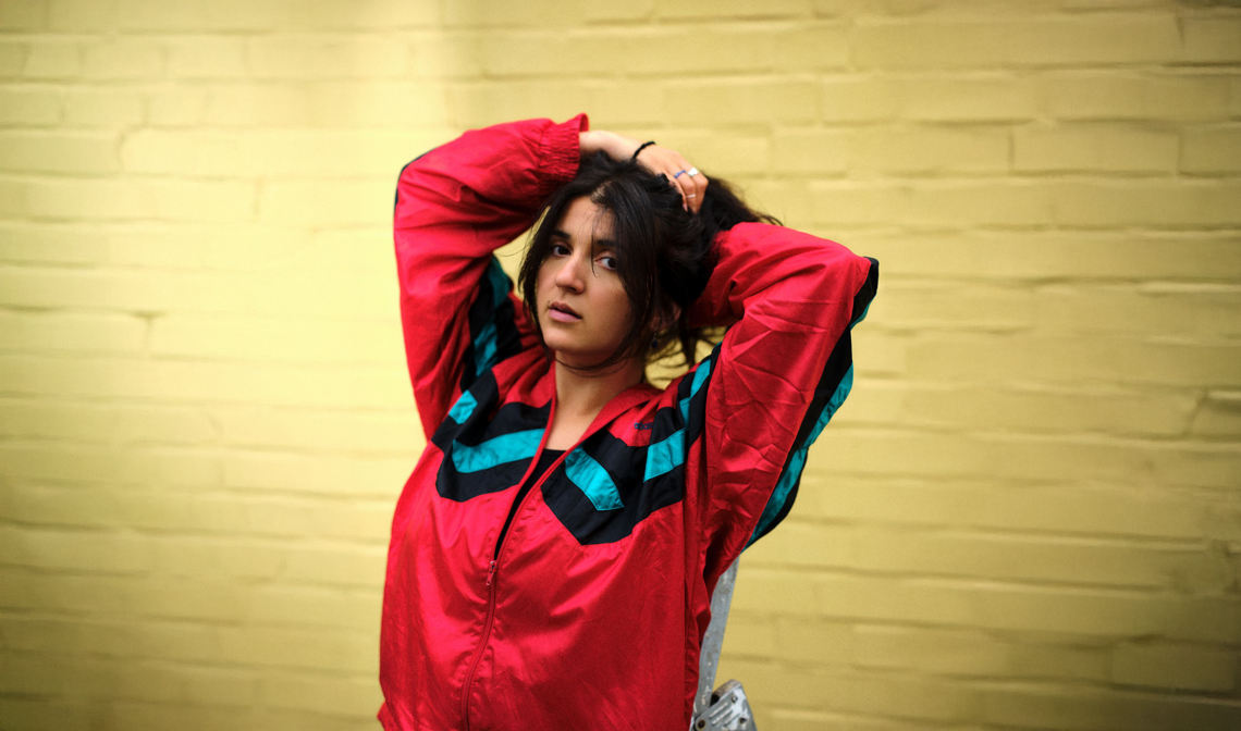 Roxana Samadi trägt eine rote Trainingsjacke mit Streifen, hält ihre Arme über den Kopf, als wolle sie ihre Haare binden. Sie steht vor einer gelben Wand.