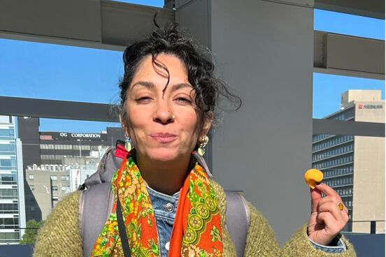Yalda Ghods trägt eine olivegrüne Wolljacke, einen Rucksack und ein buntes Tuch um den Hals. Sie lächelt mit geschlossenem Mund in die Sonne und hält einen Snack in der Hand.