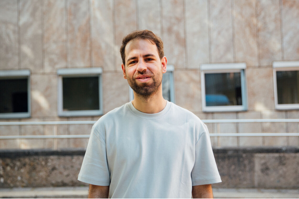 Alexander Ali Gutsfeld trägt ein helles T-Shirt und steht draußen vor einem Haus mit vielen Fenstern.